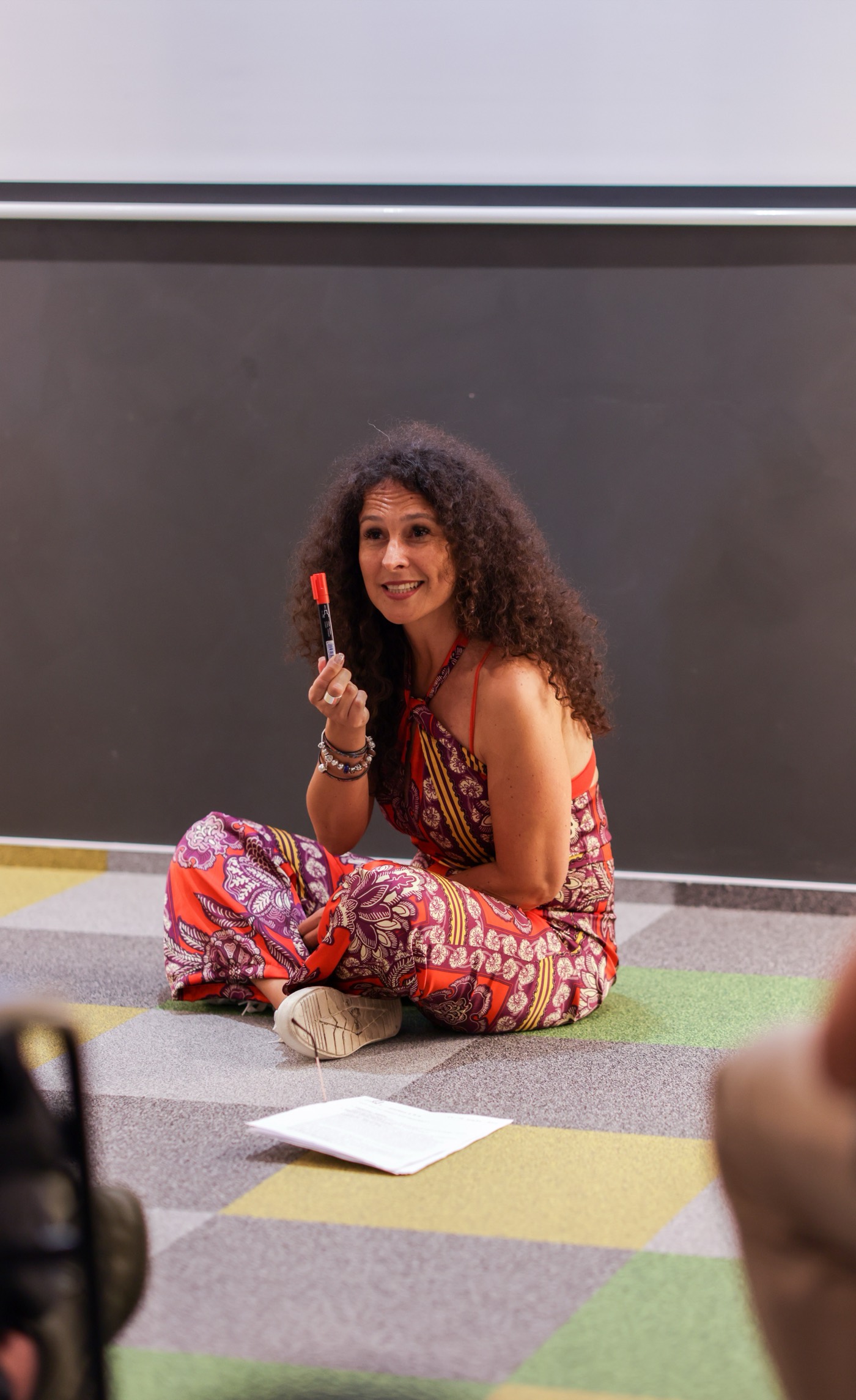 Andrea Varadi Goia sitting cross-legged on the workshop floor, marker in hand, talking to the room