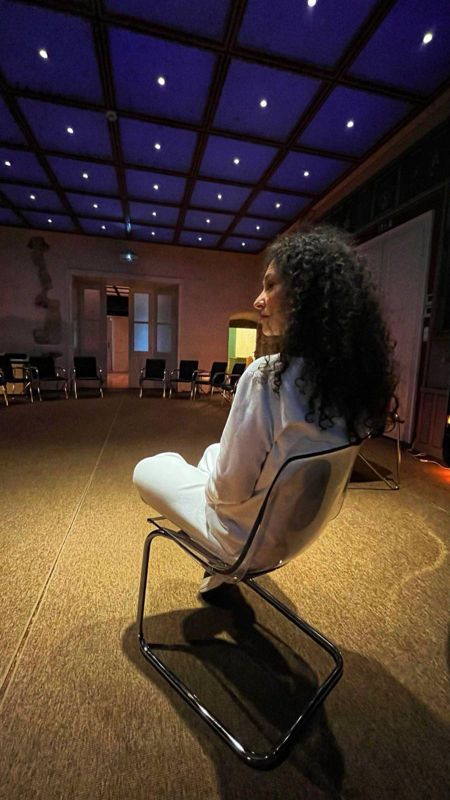 Andrea seated cross-legged in the empty workshop room, under a star-lit ceiling