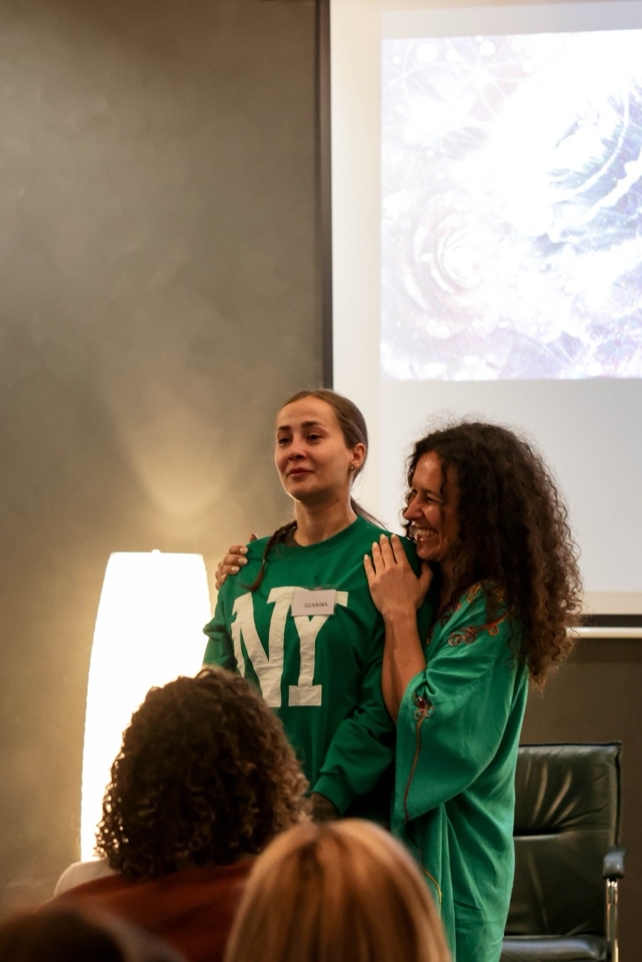 Two participants standing in front of the group, one laughing, the other steadied by a hand on her shoulder